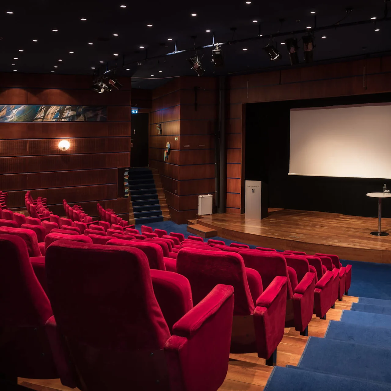 Empty red-seat auditorium faces a wooden stage with a large screen and round table holding a water bottle; wooden paneled walls with decorative lights surround the setting.