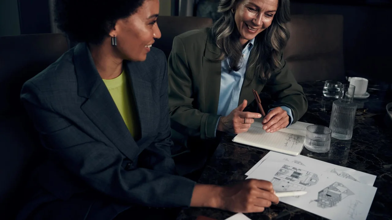 Two women reviewing documents at a table in a dimly lit room, with glasses and a carafe of water nearby.