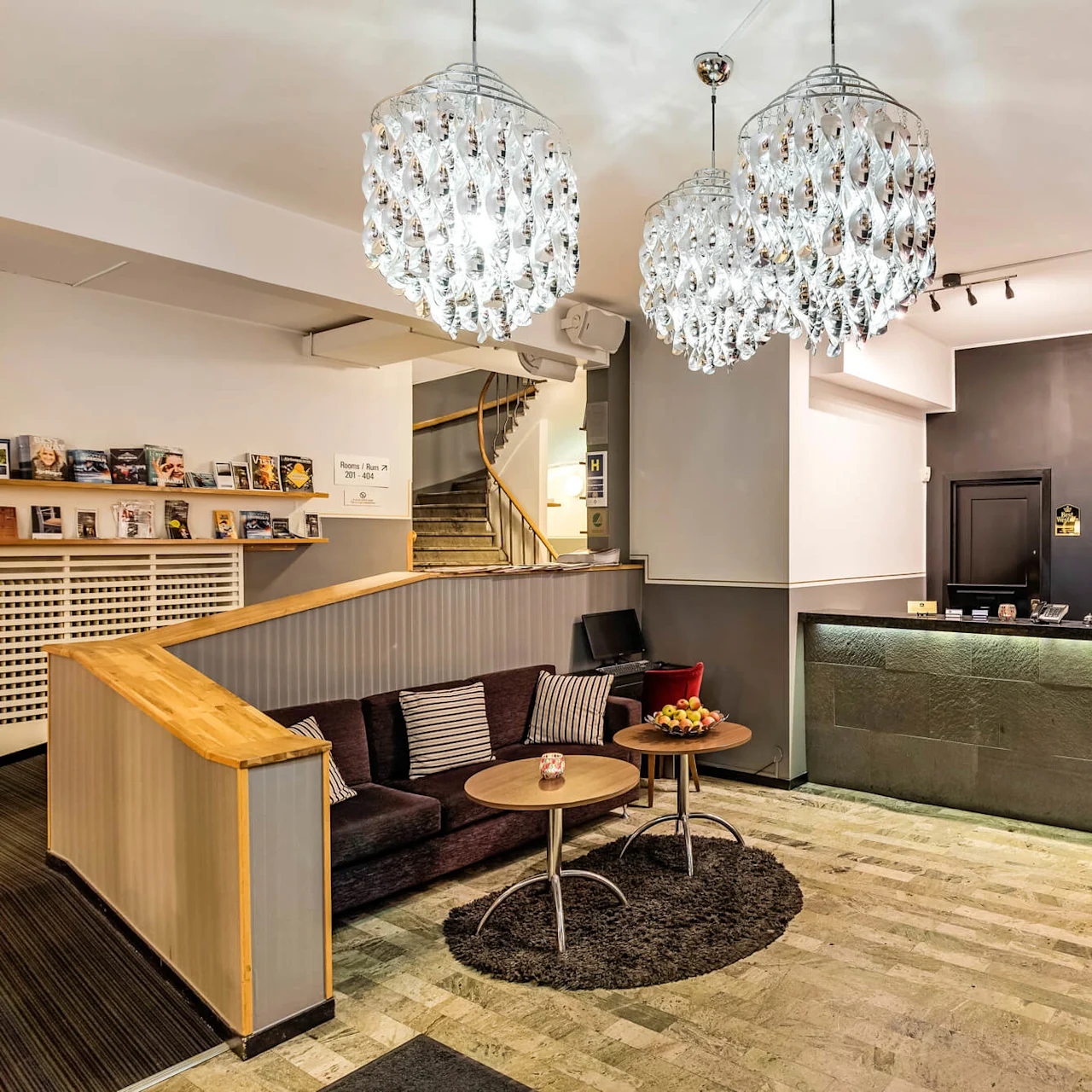 Elegant hotellresepsjon med to lysekroner, resepsjonsskranke, sitteområde, og dekorative klokker på veggen. Moderne design med en varm atmosfære.