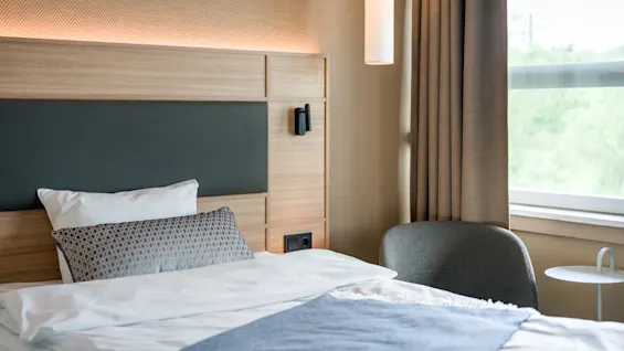A neatly made bed with white and blue bedding rests near a window. A gray chair and small round table complete the cozy, minimalist room.