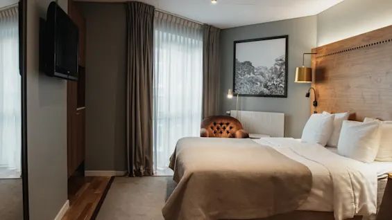 Et hyggeligt hotelværelse med en stor seng, en komfortabel lænestol og et vindue med gardiner, der skaber en afslappende atmosfære.
