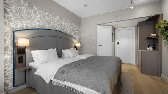 A cozy hotel room with a large bed, patterned wallpaper, and a small refreshment area. The room is ready for a comfortable stay.