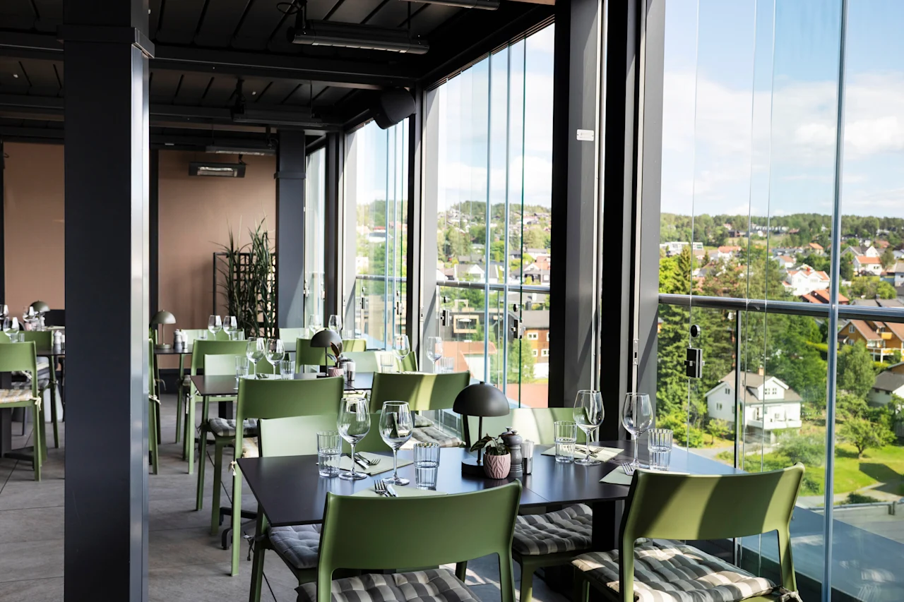 Et lyst restaurantområde med grønne stoler og mørke bord, med store vinduer som vender ut mot en by med hus og trær.