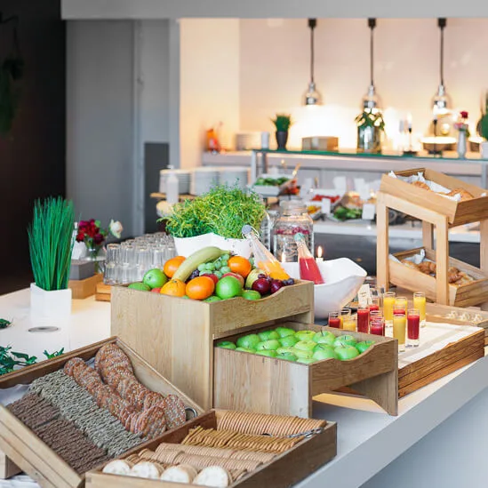 A buffet table displays fresh fruits, pastries, and juices on wooden trays in a well-lit dining area with modern decor and additional food options in the background.