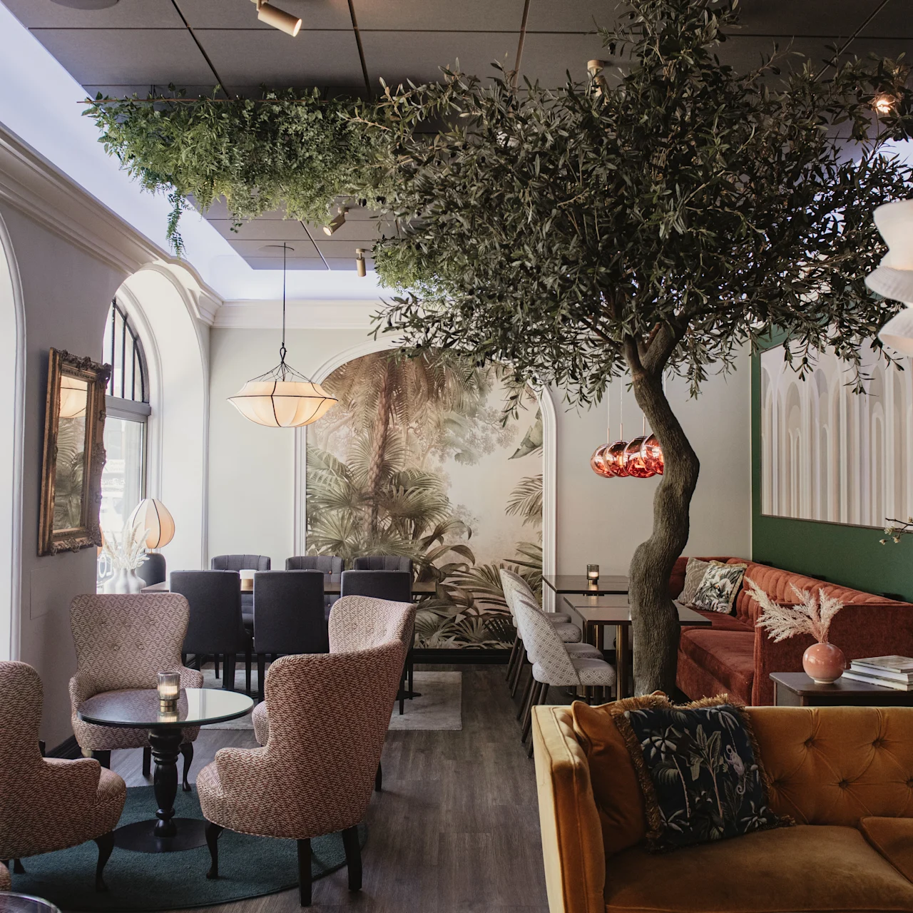Et træ udsmykker en stilfuld, hyggelig café med komfortable siddepladser, elegante lamper og store buede vinduer, der tillader naturligt lys. Et naturmotiv på væggen bidrager til den indbydende atmosfære.