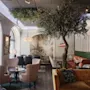 Ett träd dekorerar ett stiligt, mysigt café med bekväma sittplatser, eleganta lampor och stora välvda fönster som släpper in naturligt ljus. En muralmålning som föreställer naturen bidrar till den inbjudande atmosfären.