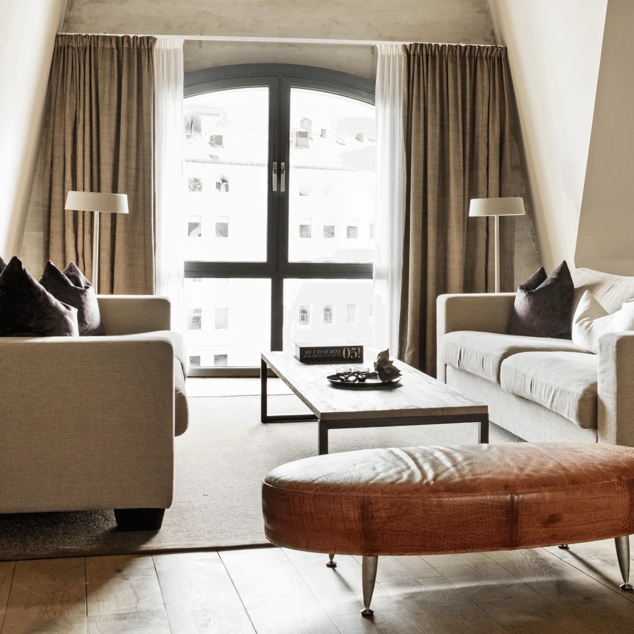 Two beige sofas face a wooden coffee table in a spacious, modern living room with a large arched window, floor lamp, and dark wooden floors. 