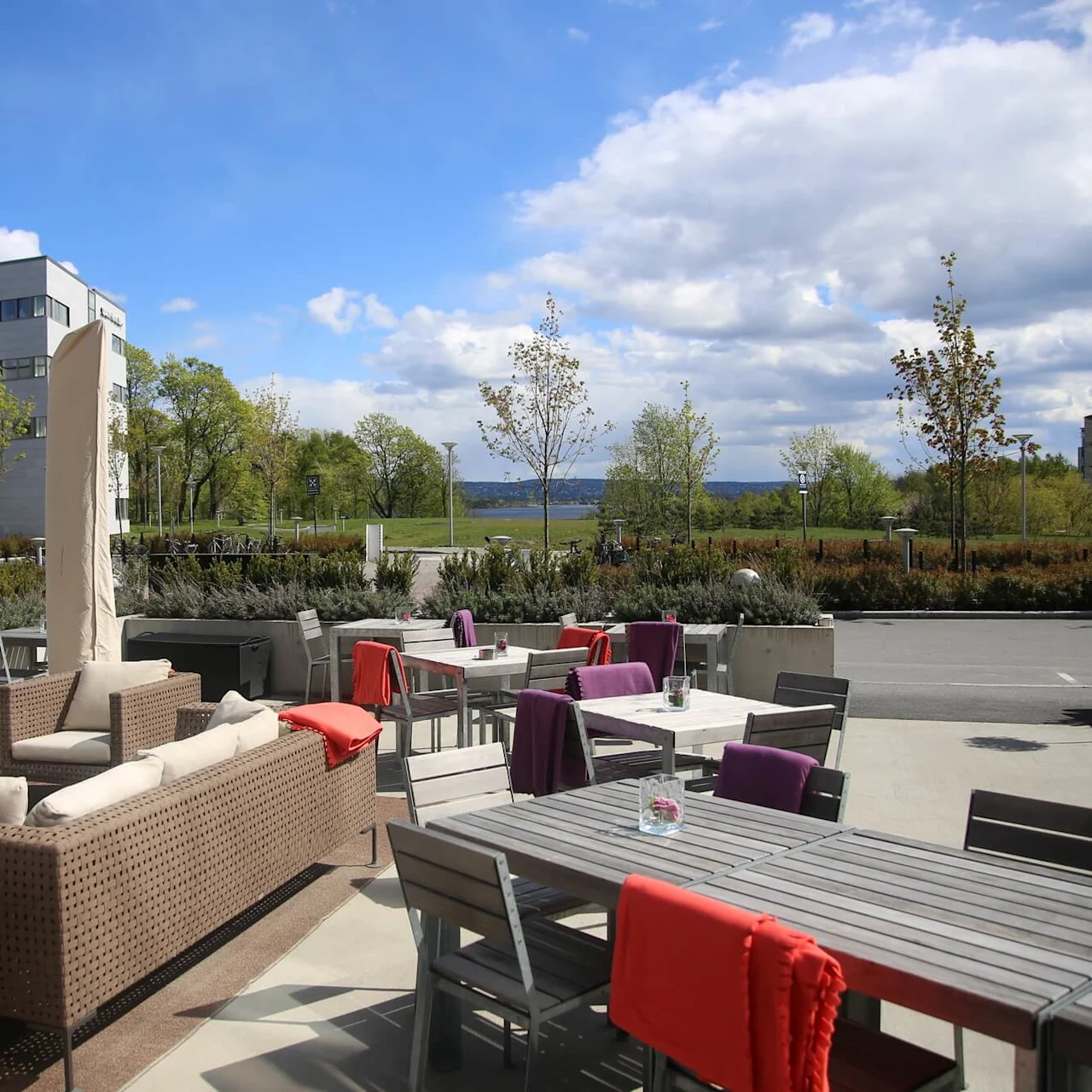 Outdoor patio with wicker sofas and tables, covered in colorful blankets. Surrounding greenery and office buildings under a partly cloudy blue sky.