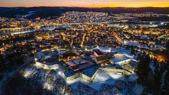 A hilltop fortress with stone walls is illuminated at dusk. It overlooks a lit cityscape with buildings, roads, and a river reflecting the sunset.