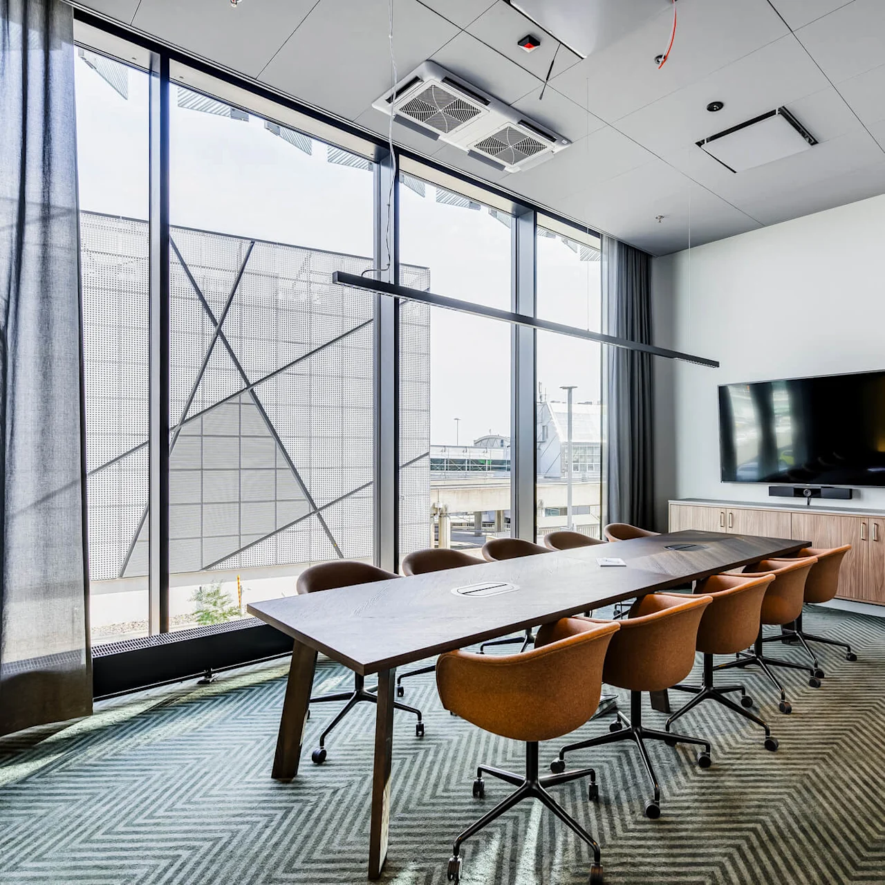A long conference table with brown chairs under a ceiling light, facing a wall-mounted TV. Large window on the left with geometric exterior view.