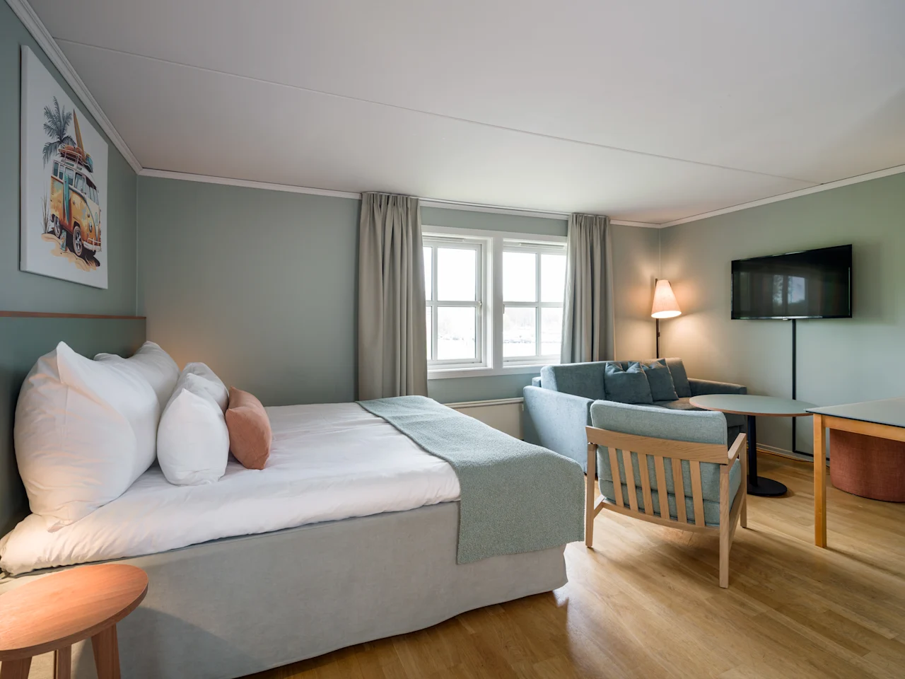 En pent redt seng med puter og teppe, ved siden av et lite bord og stol. En sofa og lampe er i bakgrunnen nær et vindu med gardiner i et koselig, mykt opplyst rom.