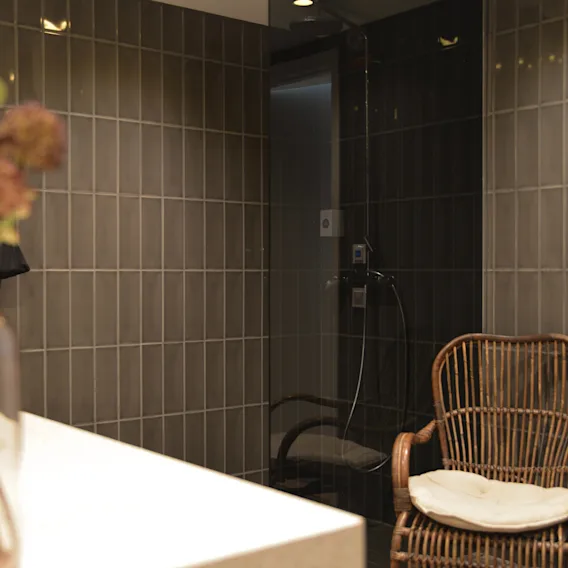 A wicker chair with a cushion sits idle in a tiled bathroom, next to a shower with dark glass doors and a countertop with a vase of flowers. 