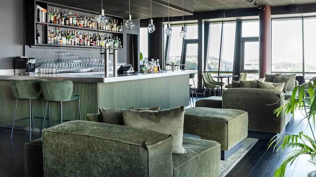 En modern bar har gröna polstrade sittplatser, en elegant disk med hängande lampor och hyllor fyllda med flaskor. Stora fönster ger utsikt över en avlägsen stadsvy.
