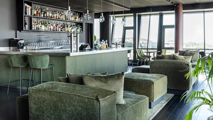 A modern bar features green upholstered seating, a sleek counter with hanging lights, and shelves stocked with bottles. Large windows provide a view of a distant cityscape.