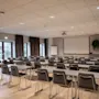 Klasserom med rekker av pulter og stoler vendt mot en tom hvit skjerm. Flippover og potteplante på siden. Naturlig lys fra vinduer med gardiner. Parkettgulv og taklamper.