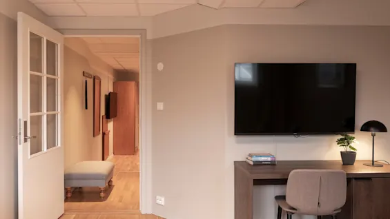 A cozy room with an open door leading to a hallway. A desk with a TV, chair, lamp, and plant suggests a comfortable workspace.