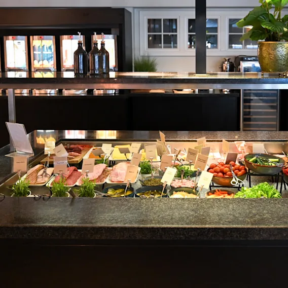 Buffet displaying various cold cuts, cheeses, and vegetables, labeled with small signs. Located in a dining area with bottles and a potted plant on a countertop above.