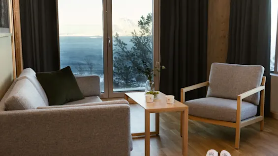 Et hyggeligt værelse med sofa, lænestol og sofabord, der byder på en fantastisk udsigt over havet og bjergene gennem et stort vindue.