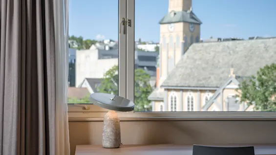 En skrivebordslampe hviler på et trebord foran et vindue med gardiner, med udsigt over en bysilhuet, der inkluderer et kirketårn under en blå himmel.
