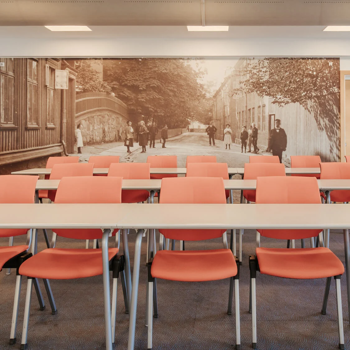 Classroom chairs and desks arranged neatly face a wall with a vintage street photograph. A plant and dispenser sit on a cabinet. An exit sign is above a door.