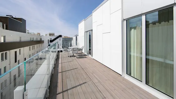 A wooden terrace features a white table and chairs, bordered by glass railings, with modern buildings visible under a clear blue sky.
