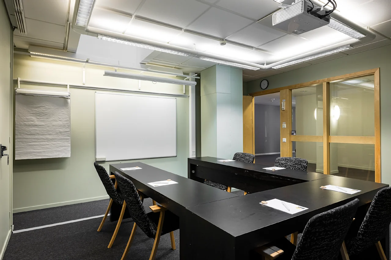 Et ryddig konferanserom med et U-formet svart bordoppsett, venter på deltakere. Stoler er pent ordnet, og notatblokker ligger på bordet. En flippover og en tavle er synlige på veggen.
