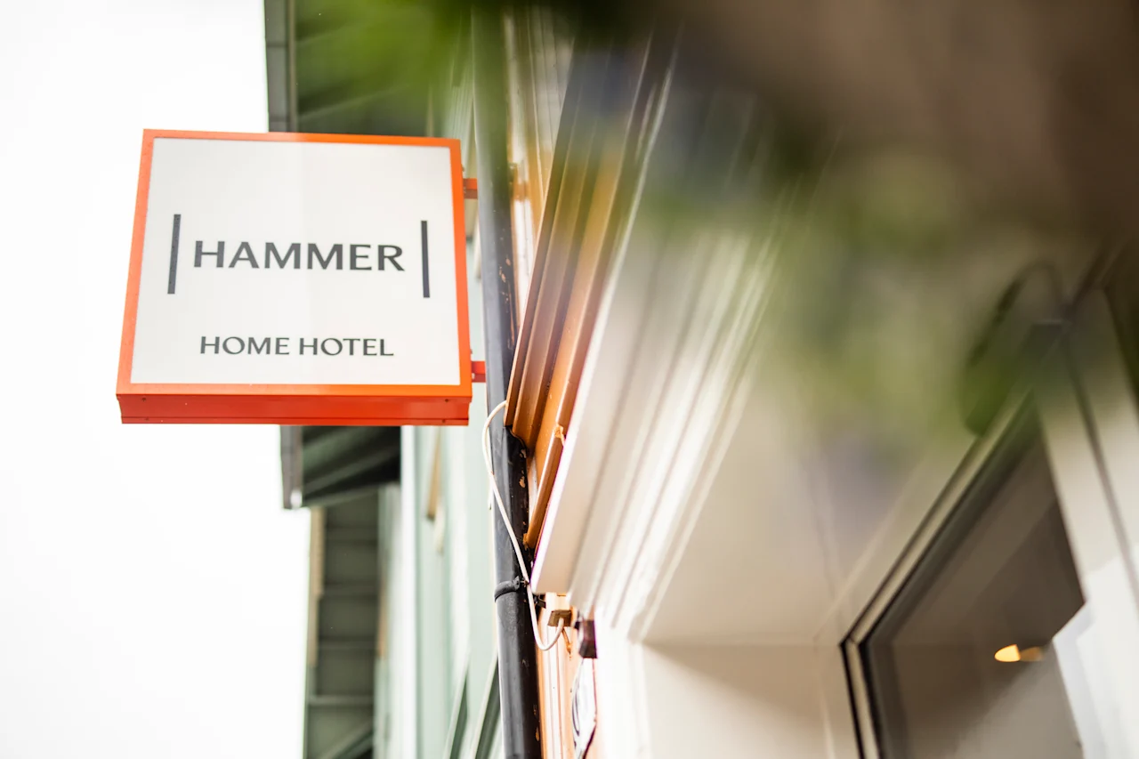A rectangular sign reads "HAMMER HOME HOTEL" in black text, bordered by orange, hanging on a building's exterior wall against a cloudy sky.