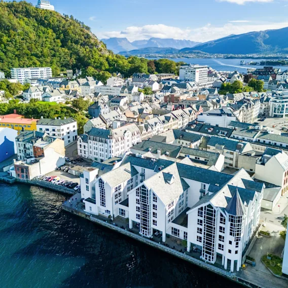 Luftfoto av Quality Hotel Ålesunds hvite sjøboder ved vannkanten, omgitt av byarkitektur og fjell.