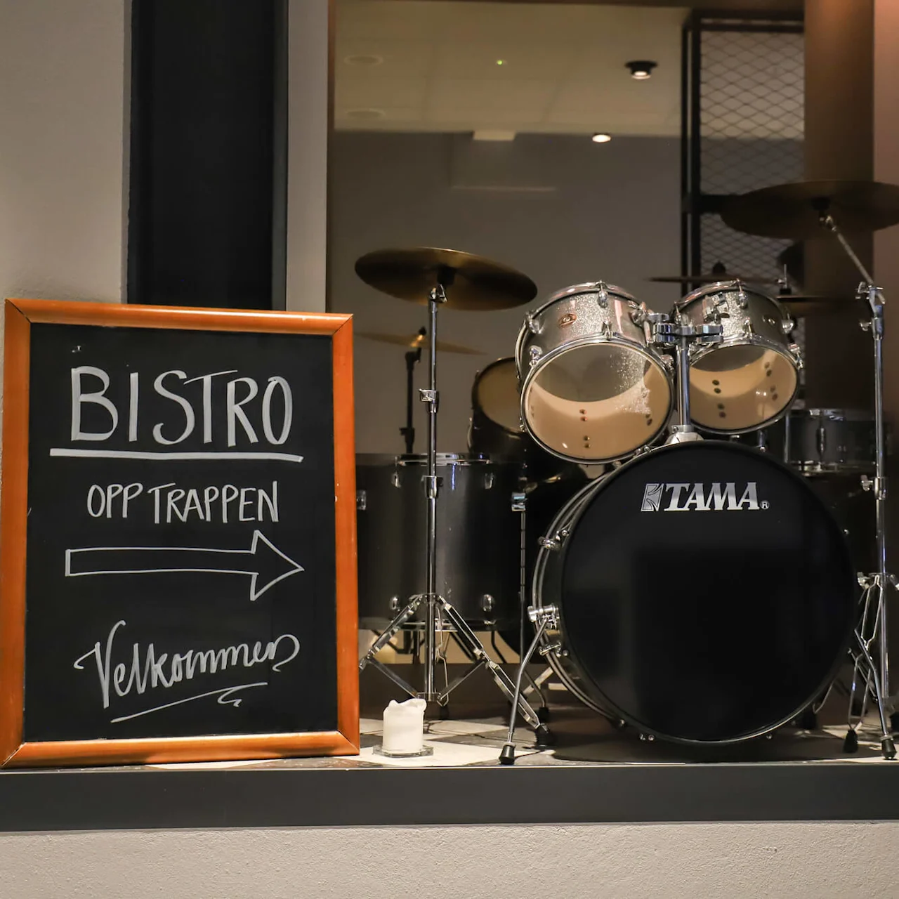 Trommesett vist på en scene ved siden av en tavle. Skiltet lyder "BISTRO OPP TRAPPEN ➔ Velkommen" i et svakt opplyst rom.