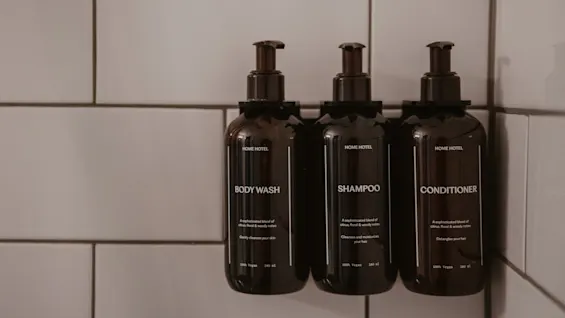 Tre mørke flasker med body wash, shampoo og balsam på en flisebelagt væg. Etiketter nævner en sofistikeret blanding og 100 procent vegansk.