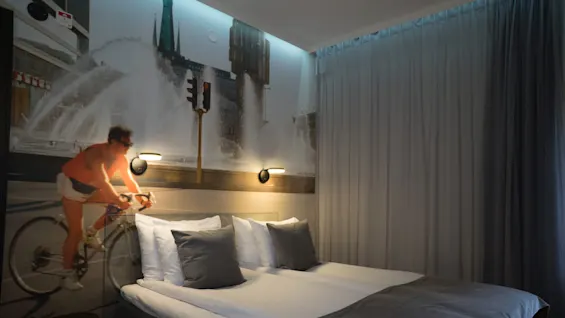 A bed with gray and white linens sits in a modern room. Behind, a large mural depicts a cyclist in front of a fountain and cityscape, with soft lighting.