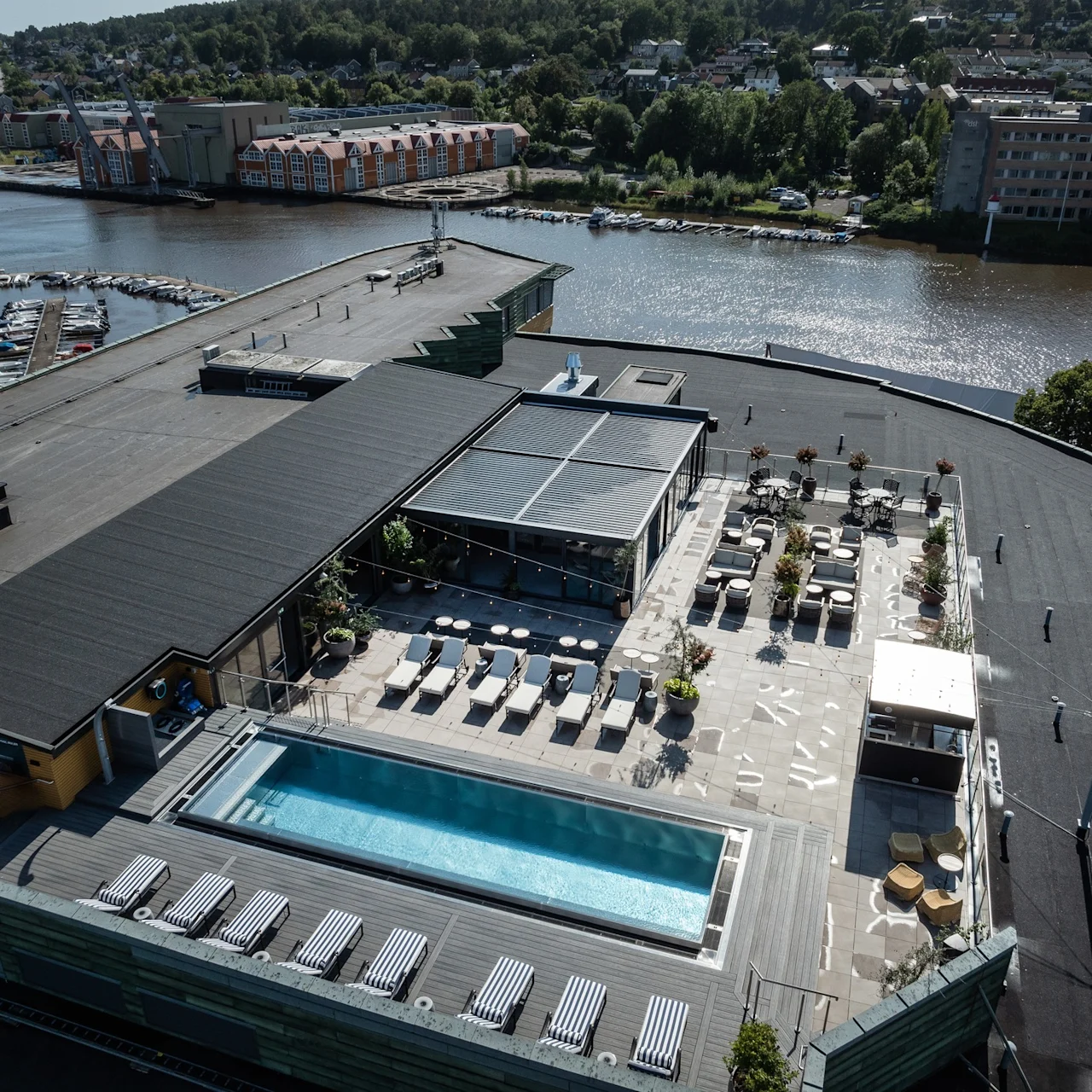 En takpool med solstolar och parasoller ligger bredvid en glasväggsbyggnad. Runtomkring finns fler sittområden och växter, med en flod och grönska i bakgrunden.