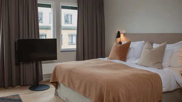 En snyggt bäddad säng med beige och bruna kuddar och filt står i ett mjukt upplyst rum. En platt-TV står framför sängen, och två fönster täcks av gardiner.