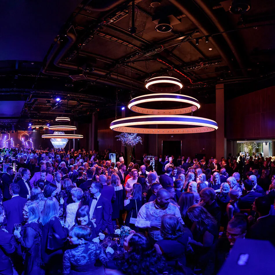 A large crowd mingles at a formal event under circular chandeliers in a dimly lit, modern venue with geometric wall patterns and ambient blue lighting.