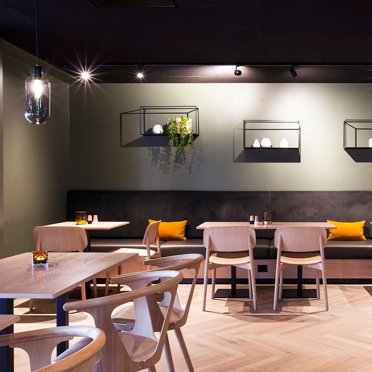 Restaurant sitteområde med trebord og stoler, en mørk benk, gule puter og dekorative hyller med planter på en grønn vegg under spotlight.