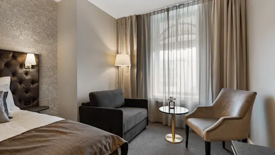 Et hyggeligt hotelværelse med en komfortabel seng, et siddeområde med sofa og lænestol, og et vindue med elegante gardiner.