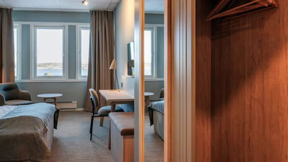 A cozy hotel room features a bed, desk, and chair. Large windows with curtains reveal a seaside view. A wooden closet with hangers is partially visible.