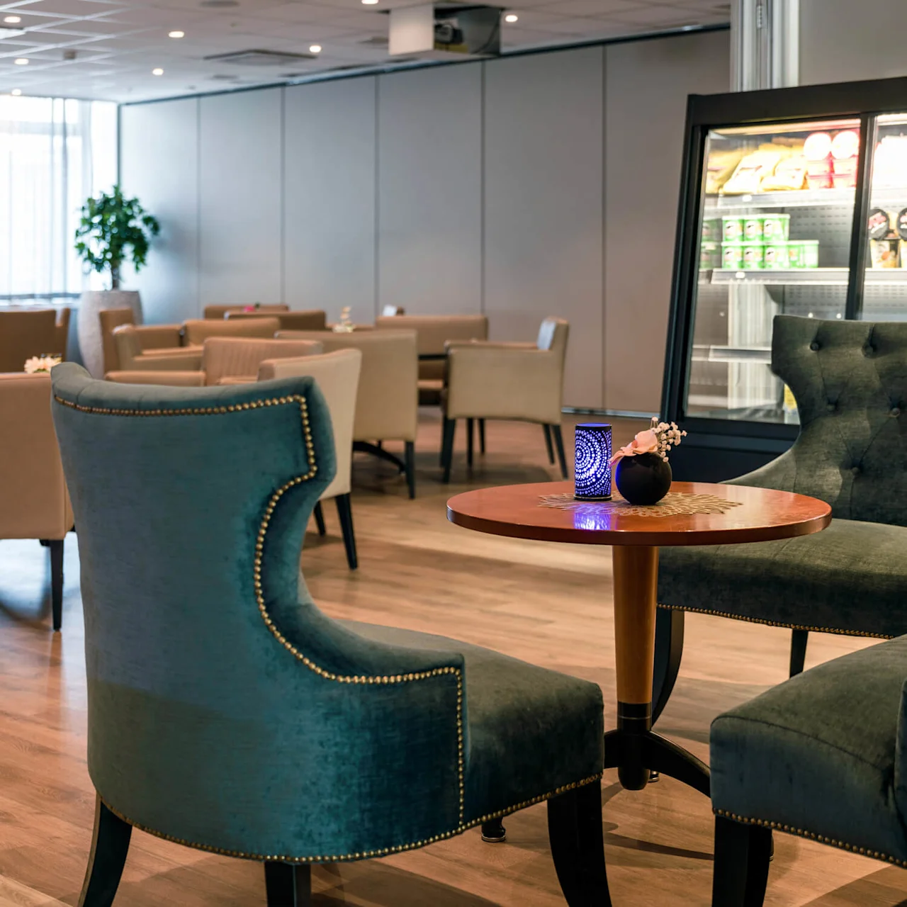 Green velvet chairs surround a wooden table with a candle in a modern lounge. Neutral armchairs are in the background near a refrigerated display, featuring snacks and drinks.