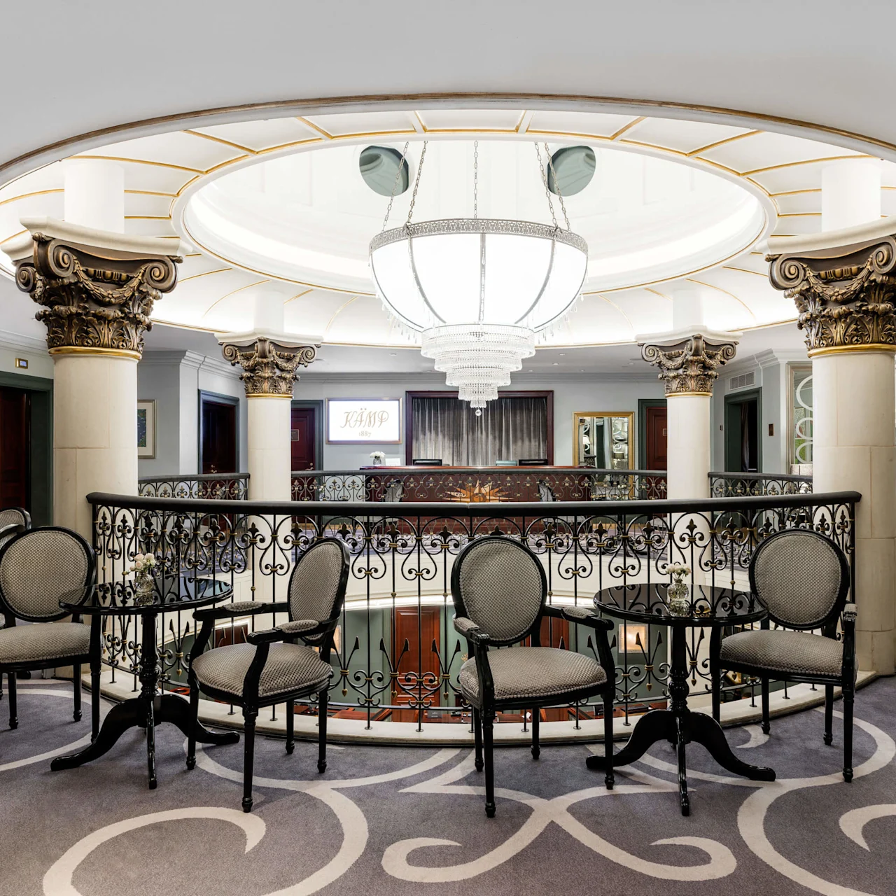 Elegant interior balcony with wrought-iron railing, surrounded by ornate columns, under a large chandelier, overlooking a sophisticated lounge area at Hotel Kämp in Helsinki.
