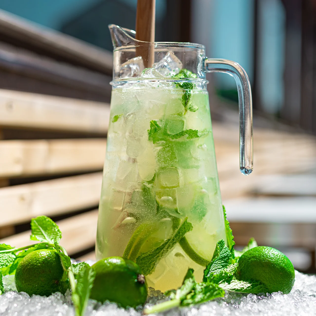 En kanna fylld med kyld mojito, mynta och lime står på ett isblock, placerat på en utomhusbänk i trä, vilket fångar en solig dagsstämning.
