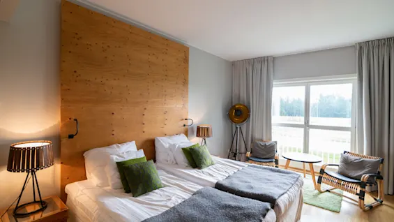 Et hyggeligt hotelværelse med to senge, en trægavl og et lyst siddeområde ved et stort vindue med udsigt over naturen.