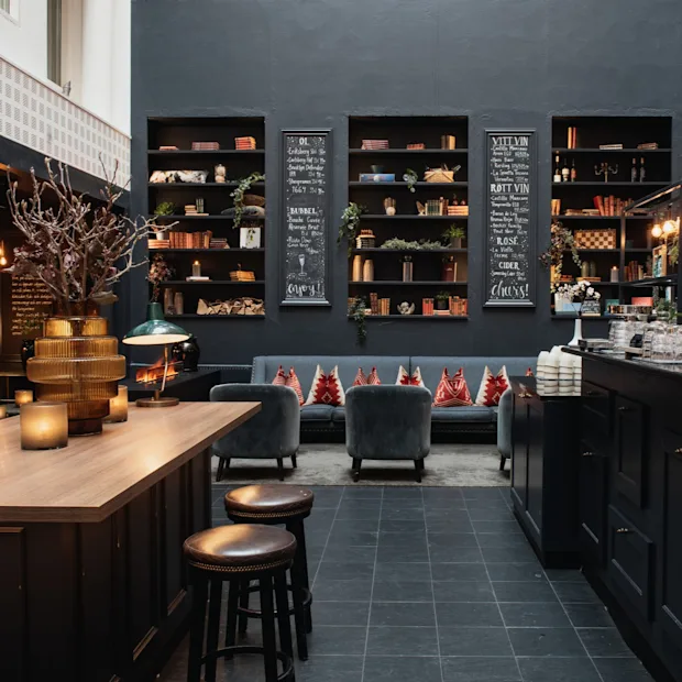 En varm og innbydende lounge med bar, komfortable sitteplasser og bokhyller. Krittavler viser OL, BUBBEL, VITT VIN, ROTT VIN, ROSE, CIDER, cheers, enjoy.