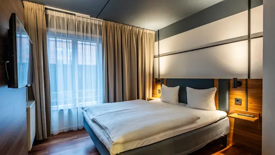 A modern hotel room featuring a double bed covered with white linens. The walls have wood paneling details, and the bed is flanked by reading lights and small bedside tables. Large curtains frame the window, allowing natural light to enter.