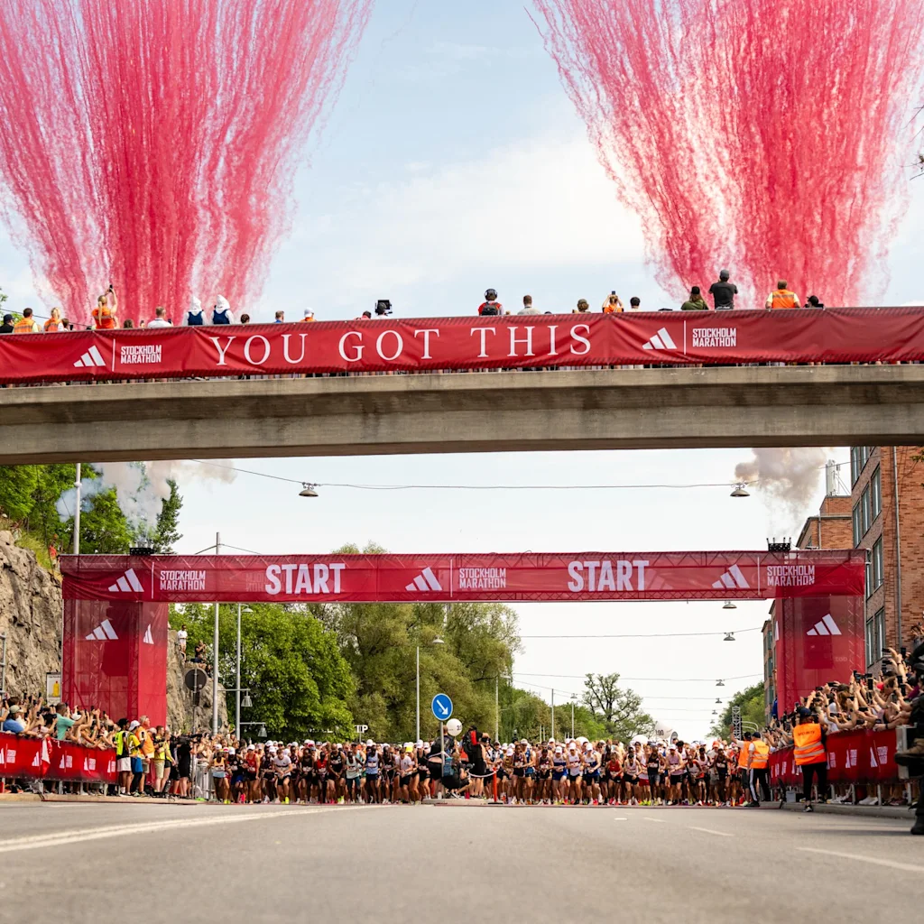 Löpare vid starten av Stockholm Marathon med röda fyrverkerier och en 'YOU GOT THIS'-banderoll ovanför.