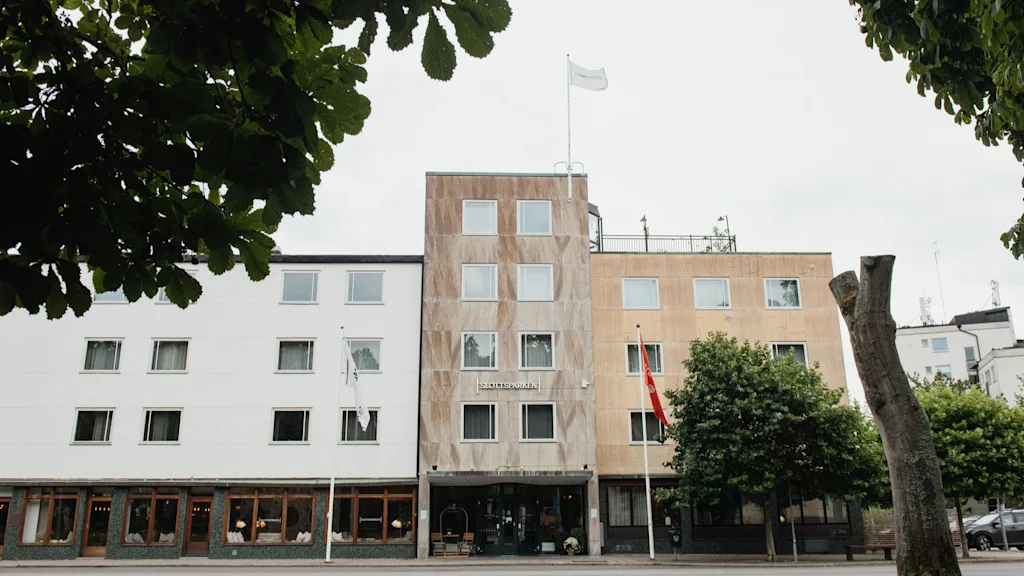 A large building with a sign reading SLOTTSSPARKEN, featuring different facades and flags, framed by green trees.