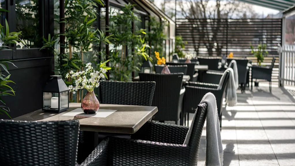 Kurvstoler og bord med vaser av blomster står under stripete markiser på en solfylt terrasse, omgitt av grøntområder og trær.