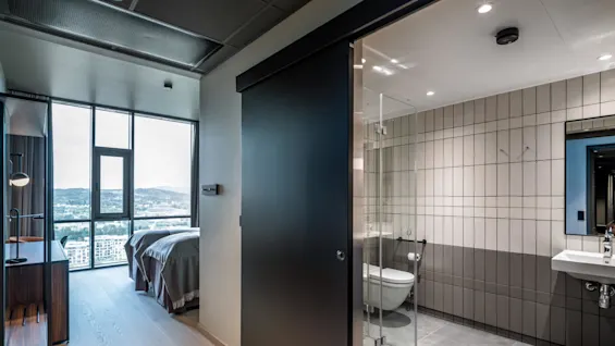 Et moderne hotellrom har et soverom med to senger og et stort vindu med utsikt over byen og fjellene, ved siden av et flislagt bad med glassdusj og hvit vask.