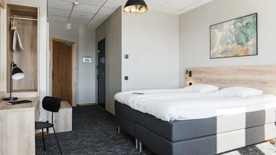 En prydligt bäddad säng med vita lakan står i ett modernt hotellrum, med en lätt träram, ett skrivbord, stol, lampa och konstverk på väggen.