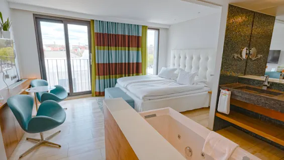 A stylish hotel room with a modern bed, colorful striped curtains, and a jacuzzi near a large window. Two blue chairs and a small table are in the corner.