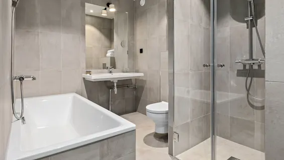 Modern badrum med badkar, dusch, handfat och toalett, alla med eleganta beslag. Neutrala kakel täcker väggar och golv, vilket skapar en ren, minimalistisk estetik.